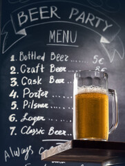 Mug of draft beer with foam on a wooden table at background of a chalk board with handwritten menu text.