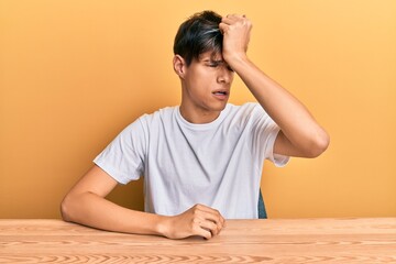 Young hispanic man wearing casual clothes sitting on the table surprised with hand on head for...