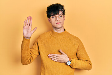 Handsome hipster young man wearing casual yellow sweater swearing with hand on chest and open palm,...