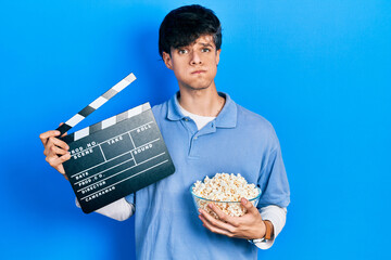 Handsome hipster young man eating popcorn holding cinema clapboard puffing cheeks with funny face....