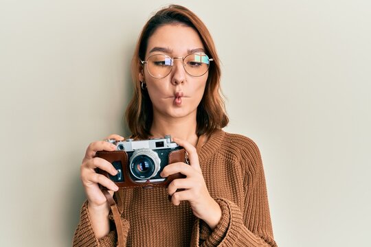 Young caucasian woman holding vintage camera making fish face with mouth and squinting eyes, crazy and comical.