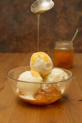 Three scoops balls of white vanilla ice cream in a bowl with apricots with yellow syrup pouring and flowing down the ice cream from a spoon on the background of a jar of jam on a wooden table