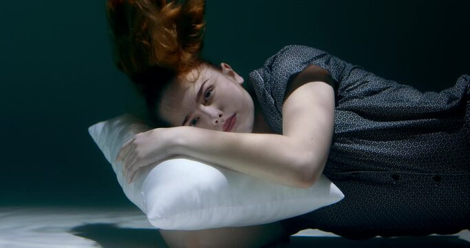 Healthy sleep. Young beautiful woman in pyjamas looks at camera smiling deep under water with head on pillow slow motion