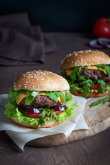 Two organic hamburger sandwiches made of grilled beef ground meat, lettuce, cheese, tomatoes and onion inside sliced bun served on cutting board on paper on dark brown wooden table. Vertical image