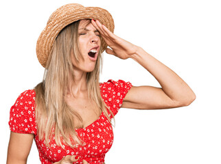 Beautiful caucasian woman wearing summer hat surprised with hand on head for mistake, remember...