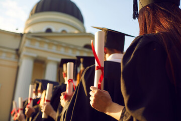 Students on university graduation ceremony day. Rare view on graduates standing in line holding degree diploma against blue sky and educational institution building on background. Education concept