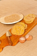 Top view, medium distance of a wood cutting board with a knife and three slices of peeled eggplant  with breadcrumbs, a paper plate of breadcrumbs, and two eggs egg shells, all on a brown burlap cloth