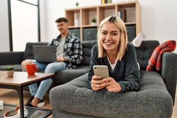 Obraz premium Young caucasian couple smiling happy using laptop and smartphone sitting on the sofa at home.