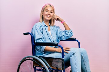 Beautiful blonde woman sitting on wheelchair smiling pointing to head with one finger, great idea...