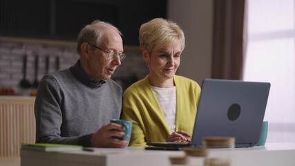 two retirees are using laptop at home kitchen, surfing internet, shopping online, old man and woman are viewing pictures on display