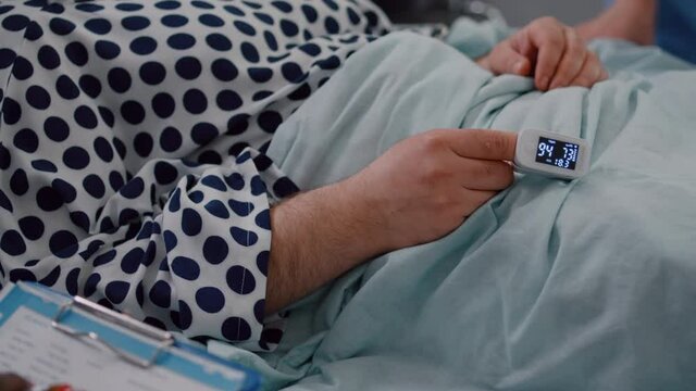 Closeup of sick man patient resting in bed with medical oximeter on finger. Surgeon doctor checking heart pluse analyzing illness cardiogram working in hospital ward explaining disease diagnosis