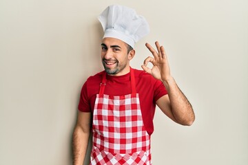 Young hispanic man wearing baker uniform smiling positive doing ok sign with hand and fingers....