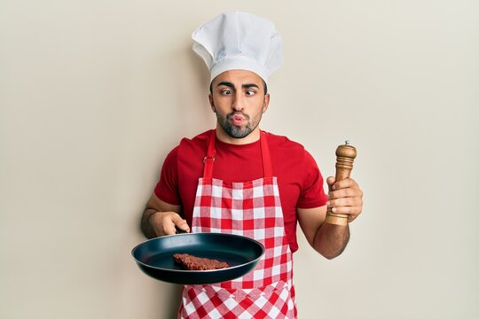 Young hispanic man wearing professional cook uniform and hat cooking beef making fish face with mouth and squinting eyes, crazy and comical.