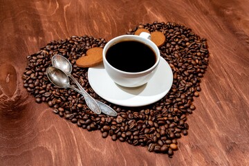 White coffee mug, heart shaped gingerbread and two spoons on a heart shaped base made from coffee beans