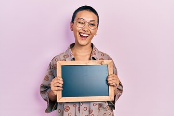 Beautiful hispanic woman with short hair holding blackboard winking looking at the camera with sexy...