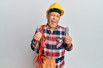 Senior hispanic man wearing handyman uniform holding led lightbulb and incandescent bulb sticking tongue out happy with funny expression.