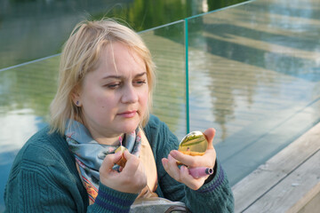 Middle-aged woman with blond hair looking in mirror and wears lipstick with brush while sitting on bench in a city park. The woman freshen her makeup.