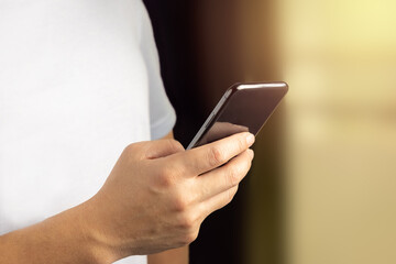 A man person hold smartphone (mobile phone) and using it by look into its screen while searching for information or playing social media or Internet. Orange sunlight in the background. White shirt.