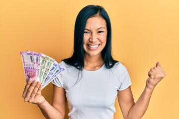 Beautiful hispanic woman holding indian rupee banknotes screaming proud, celebrating victory and...