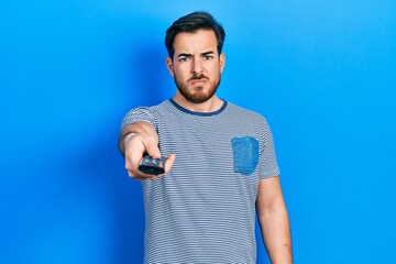 Handsome caucasian man with beard holding television remote control skeptic and nervous, frowning upset because of problem. negative person.