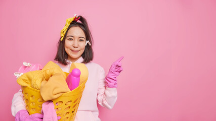 Obraz na płótnie Canvas Pleased Asian woman housekeeper with dark hair wears sweatshirt and protective rubber gloves holds laundry basket indicates aside on blank space isolated over pink background. Housecleaning concept