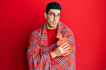 Handsome hispanic man wrapped in a winter blanket holding hot water bag smiling looking to the side and staring away thinking.