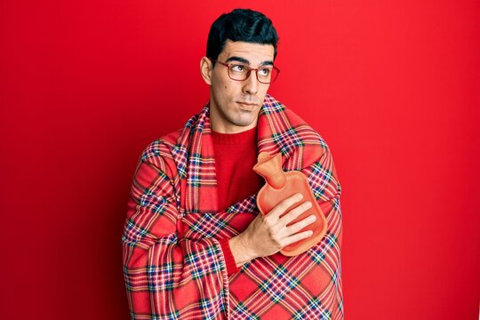 Handsome Hispanic Man Wrapped In A Winter Blanket Holding Hot Water Bag Smiling Looking To The Side And Staring Away Thinking.