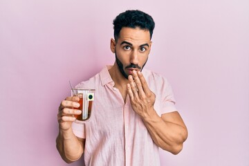 Young hispanic man holding cup of tea covering mouth with hand, shocked and afraid for mistake....