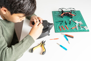 Concentrated boy repairing the remote control of his toy drone with a screwdriver in his hand and various tools and spare parts on the table for his drone.