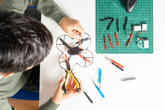 View from above of concentrated boy repairing the elices of his toy drone with a clamp in his hand and various tools and spare components on the table for his drone.