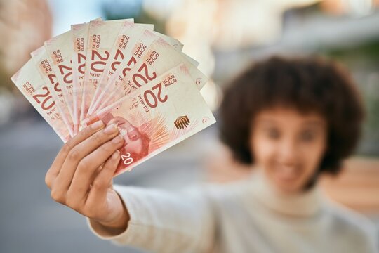 Young hispanic girl smiling happy holding israel shekels at the city.