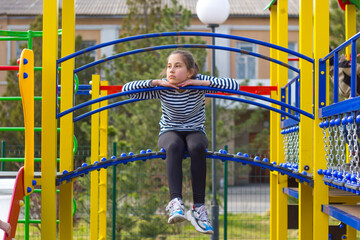 Brunette girl 10 years old, bored on the playground, sits on a slide with her legs dangling. 