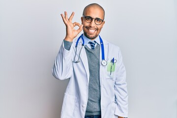 Hispanic adult man wearing doctor uniform and stethoscope smiling positive doing ok sign with hand and fingers. successful expression.