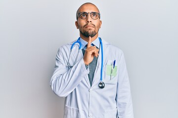 Hispanic adult man wearing doctor uniform and stethoscope thinking concentrated about doubt with finger on chin and looking up wondering