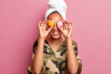Pleased smiling young woman covers sweet macaroons over eyes being in good mood wears dressing gown...