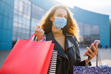 Smiling woman in protective medical mask with shopping bags using her phone. Young woman after shopping on the city street. Online shopping concept. Shopping in the coronavirus epidemic. 