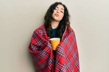 Fototapeta premium Young brunette woman with curly hair wrapped in a red warm red blanket drinking coffee looking at the camera blowing a kiss being lovely and sexy. love expression.