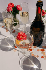 Crystal coupe with champagne with a pink flower in it, and a bottle, on table in sun light.