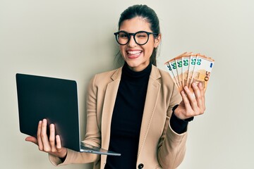 Young hispanic woman wearing business style holding laptop and 50 euros winking looking at the...