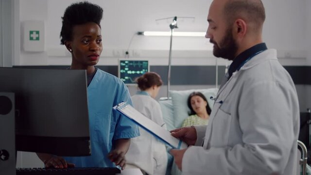 Black assistant typing health care treatment on computer while in background surgeon doctors checking sick woman writing illness diagnosis. Patient lying in hospital ward bed during consultation