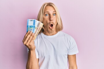 Caucasian young man with long hair holding hungarian forint banknotes scared and amazed with open mouth for surprise, disbelief face