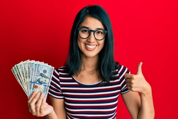 Beautiful asian young woman holding dollars smiling happy and positive, thumb up doing excellent and approval sign