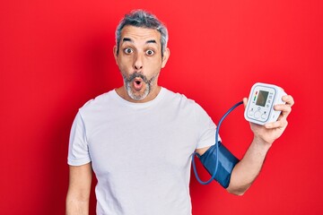 Handsome middle age man with grey hair using blood pressure monitor scared and amazed with open...