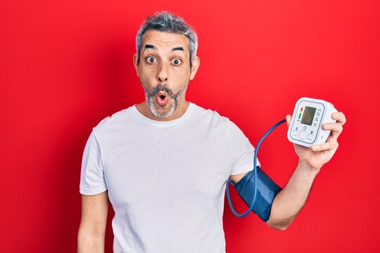 Handsome middle age man with grey hair using blood pressure monitor scared and amazed with open mouth for surprise, disbelief face