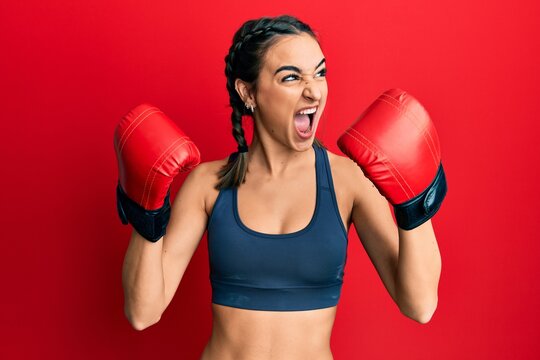 Young brunette girl using boxing gloves wearing braids angry and mad screaming frustrated and furious, shouting with anger. rage and aggressive concept.