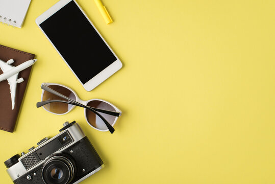 Top view photo of smartphone display notebook pen sunglasses camera and plane model on passport cover on isolated pastel yellow background with copyspace