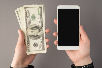 First person top view photo of woman's hands holding smartphone and stack of money hundred dollar banknotes on isolated grey background with copyspace