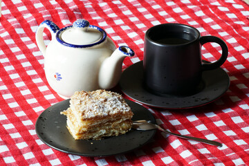 A piece of cake (pie), a cup of tea and a teapot on a bright red and white checkered tablecloth. A small treat for tea or a light breakfast in a cafe.
