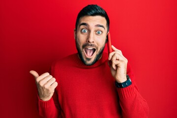 Young hispanic man having conversation talking on the smartphone pointing thumb up to the side...