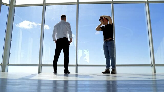 Male photographer taking photos of a businessman. Rear view of a young man in white shirt and black trousers posing on camera indoors. Panoramic windows.